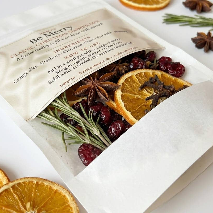 A white stand-up pouch labeled 'Be Merry' with dried orange slices, cranberries, rosemary, and star anise visible through a clear window, styled on a white surface with dried orange slices and star anise around it.